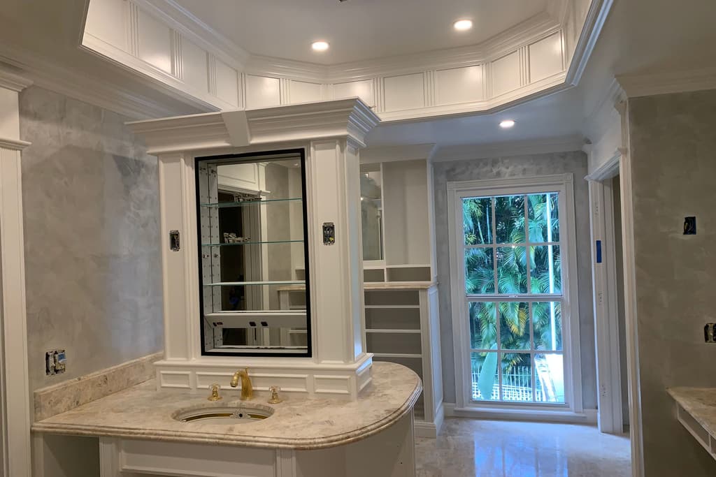 A white vanity with a curved marble top, gold fixtures and a black-framed mirrored cabinet, with a tall window to palms beyond.