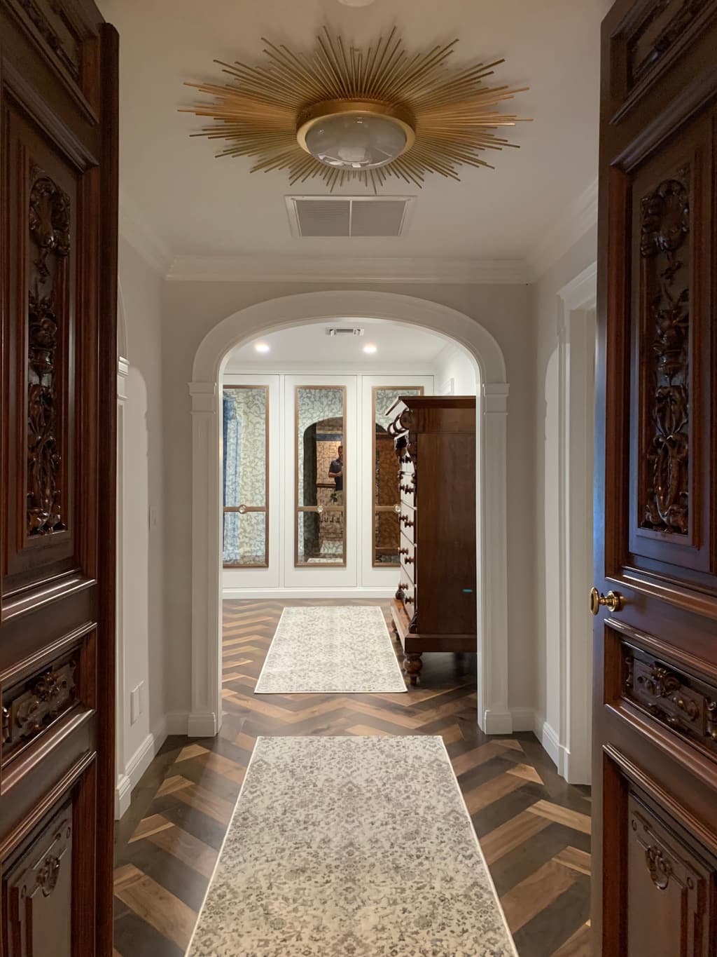 Carved wood double doors stand open onto a white hallway with a gold sunburst ceiling light, an arched opening and runners on a herringbone wood floor.